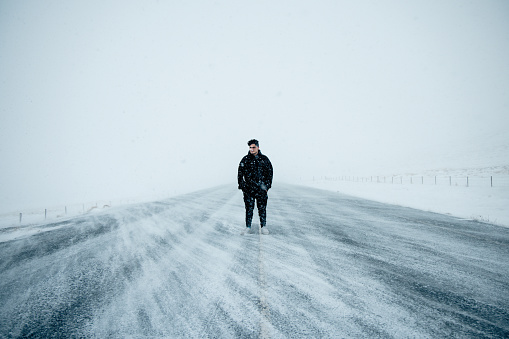 Young man standing in the middle of snowcovered icelandic country road, hands in pocket looking over the the snow covered highlands. Extreme windy snow weather, snowflakes flying over the highlands road. Icelandic Highjlands in Winter. Young Traveller Road Trip Portrait. Highland Iceland, Northern Europe, Nordic Countries.