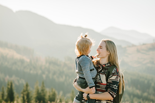Mother with kid hiking in autumn mountains. Young tourists on top of a mountain enjoying sunset. Happy family. Mom with backpack hugging daughter. Holiday trip concept. World Tourism Day. Back view.