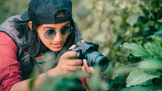 In this outdoor day image, an Asian/Indian confident young female photographer shoots macro-images of nature. The photographer is wearing a black cap, a warm sleeveless jacket with big pockets, and round sunglasses. She shoots in the hills of Himachal Pradesh.