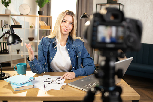 Happy blond caucasian woman in jeans shirt recording online tutorial on modern camera. Smiling female using laptop and camera during remote work at home.