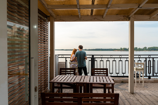 Rear view of unrecognizable young couple embracing at fence near the river at terrace of cozy cafe