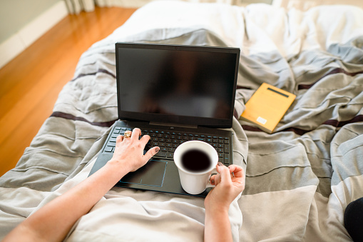POV of a woman drinking coffee and working on a laptop while sitting up in her bed in the morning