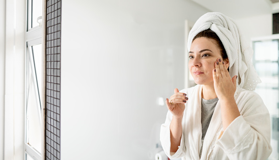 Woman with a towel wrapped around her hair and wearing a bathrobe putting cream on her face in her bathroom
