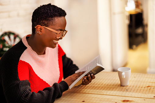A pretty young female black woman is reading a book while resting for table in the dining room Scandinavian-style living room in an apartment. Domestic life. Selective focus.