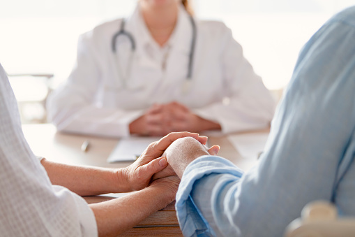 Mature couple holding hands at a doctors office. Doctor can be seen in the background.