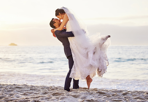 Romantic wedding couple barefoot on pebblestone beach at river on late summer afternoon, groom carrying bride, smiling at each other, full length, copy space