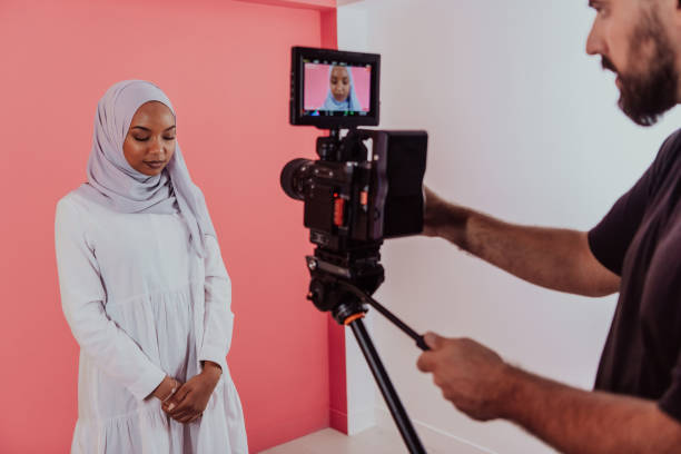 istock Videographer in digital studio recording video on professional camera by shooting female Muslim woman wearing hijab scarf plastic pink background 1390167113