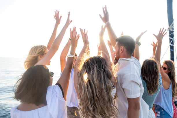 Group of people with raised hands standing on yacht deck. Having good time at sunset. Summer vacation, youth and fun concept.