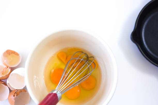 Eggs in Bowl with Whisk, Eggshells, Frying Pan, White Background stock photo