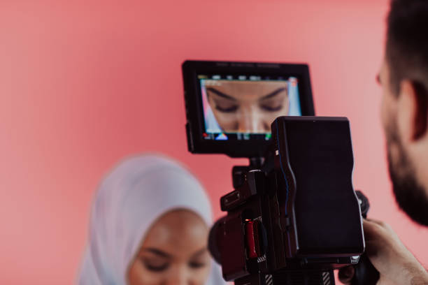 istock Videographer in digital studio recording video on professional camera by shooting female Muslim woman wearing hijab scarf plastic pink background 1390175414