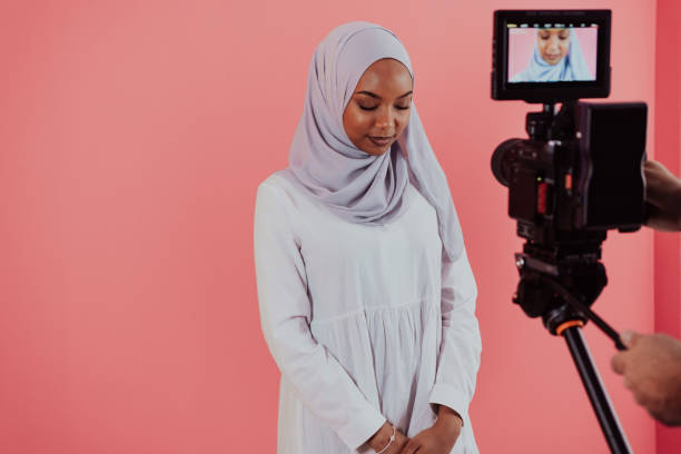 istock Videographer in digital studio recording video on professional camera by shooting female Muslim woman wearing hijab scarf plastic pink background 1390183793