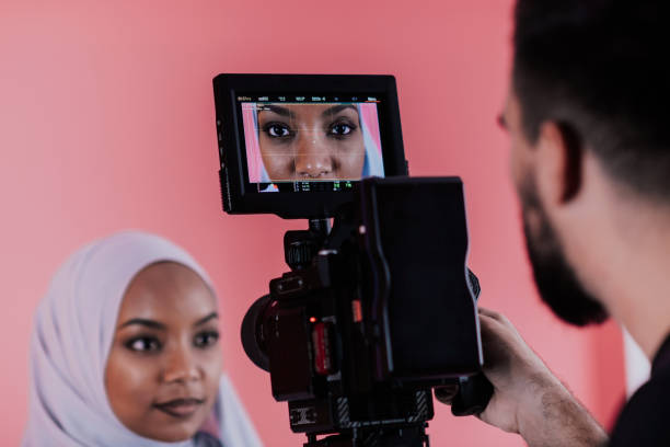 istock Videographer in digital studio recording video on professional camera by shooting female Muslim woman wearing hijab scarf plastic pink background 1390188218