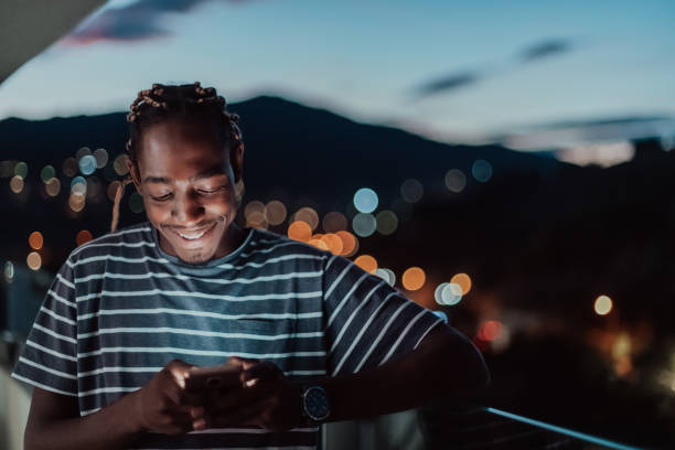 el joven en una calle urbana de la ciudad por la noche enviando mensajes de texto en un teléfono inteligente con bokeh y luces de neón de la ciudad en el fondo - typebar fotografías e imágenes de stock