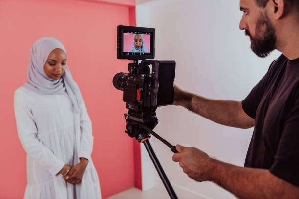 istock Videographer in digital studio recording video on professional camera by shooting female Muslim woman wearing hijab scarf plastic pink background 1390205335