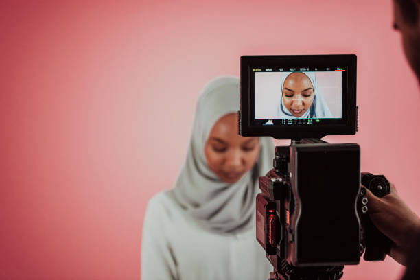 istock Videographer in digital studio recording video on professional camera by shooting female Muslim woman wearing hijab scarf plastic pink background 1390209392
