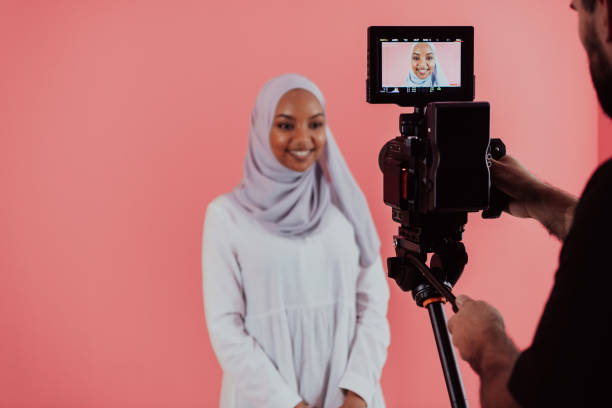 istock Videographer in digital studio recording video on professional camera by shooting female Muslim woman wearing hijab scarf plastic pink background 1390209615