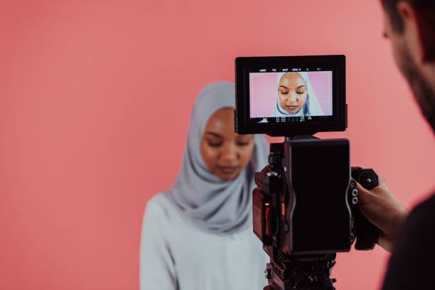 istock Videographer in digital studio recording video on professional camera by shooting female Muslim woman wearing hijab scarf plastic pink background 1390210544