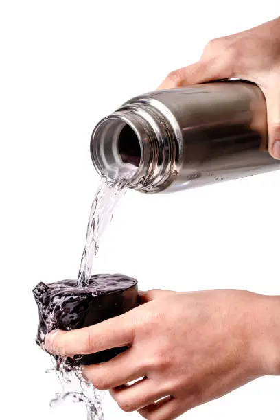 The hand was holding the stainless water bottle to pour water into the black lid and overflow isolated on a white background.