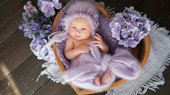 Newborn girl dressed in woolen cap and garment lies in heart-shaped wooden basket among purple flowers. Photo-shoot of cute baby in studio close upper view