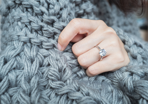 Close up of an elegant engagement diamond ring on woman finger with dark gray sweater winter clothe. love and wedding concept. Soft and selective focus.