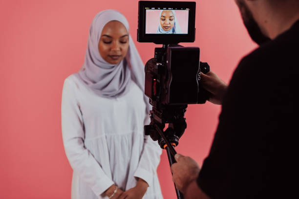 istock Videographer in digital studio recording video on professional camera by shooting female Muslim woman wearing hijab scarf plastic pink background 1390214894
