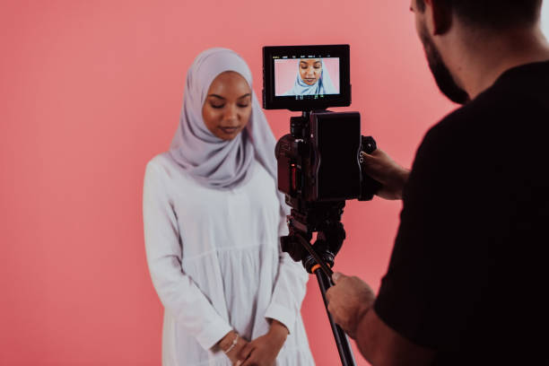 istock Videographer in digital studio recording video on professional camera by shooting female Muslim woman wearing hijab scarf plastic pink background 1390215272
