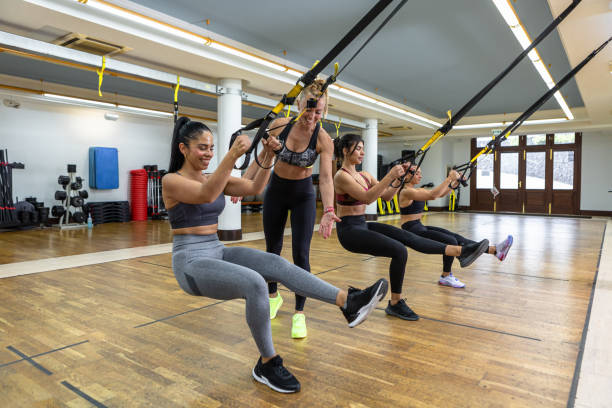 Sportswomen exercising with TRX ropes together Positive female coach smiling and watching athletes squatting with raised legs during suspension training in spacious gym suspension training stock pictures, royalty-free photos & images
