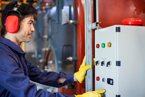 Close up side view of mid adult Asian American industrial worker standing in front of a control panel with buttons and switches