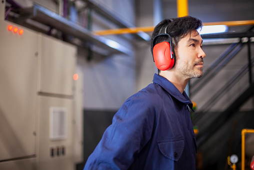 Side view of a mid adult Asian American male industrial worker wearing ear muffs standing inside an industrial facility