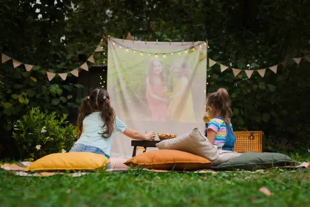 Photo of Backyard Family outdoor movie night with kids. Sisters spending time together and watching cimema at backyard. DIY Screen with film. Summer outdoor weekend activities with children. Open air cinema.