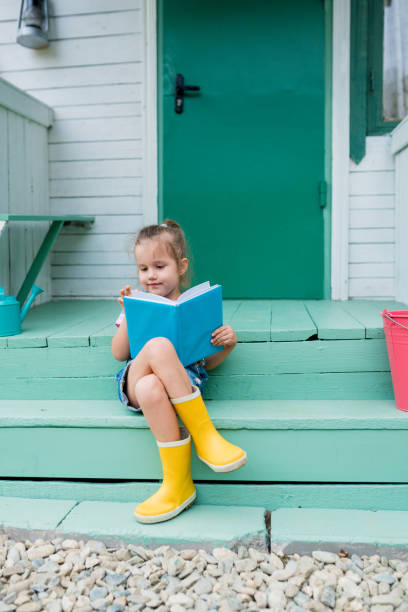 urocza dziewczynka siedząca na schodach i czytająca książkę na podwórku. maluch rozwija samodzielne czytanie na zewnątrz. wieś - book child staircase steps zdjęcia i obrazy z banku zdjęć