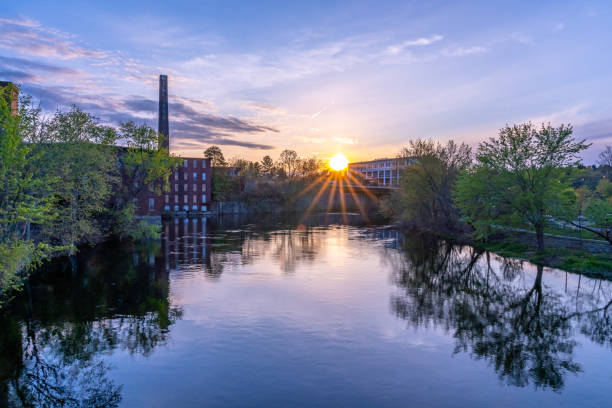The sun and its rays at sunset with reflection on the Nashua River. On the shore there is a historical building of a cotton factory with a tall brick chimney in an old industrial park The sun and its rays at sunset with reflection on the Nashua River. On the shore there is a historical building of a cotton factory with a tall brick chimney in an old industrial park. Nashua, NH nashua new hampshire stock pictures, royalty-free photos & images