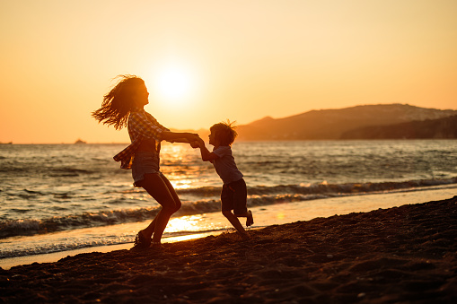 Happy mother having fun while holding hands with her small boy and spinning on the beach at sunset. Copy space.