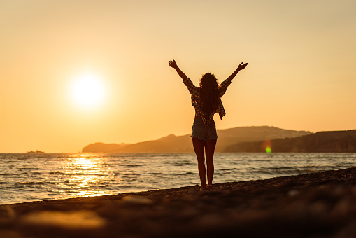 Rear view of carefree woman standing on the beach with raised arms at sunset. Copy space.