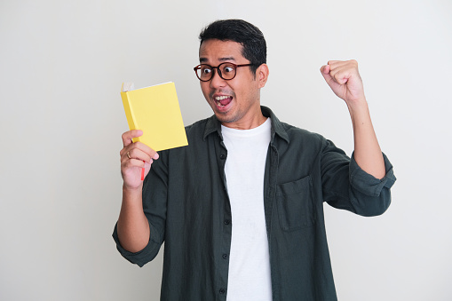 Adult Asian man showing excited gesture with fist clenched when reading a book