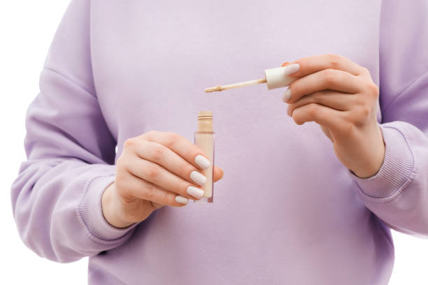 Close up shot of a woman in a lavander oversized sweatshirt holding a concealer bottle and a brush in hands. Nice groomed nails with nail polish. Close up shot of a woman in a lavander oversized sweatshirt holding a concealer bottle and a brush in hands. Nice groomed nails with nail polish. concealer stock pictures, royalty-free photos & images