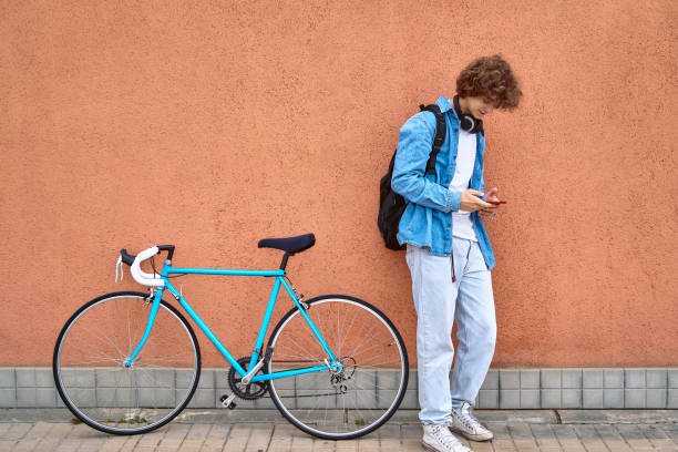 istock Young curly-haired student with headphones standing on wall outdoors using cell phone next to her bicycle 1390251699