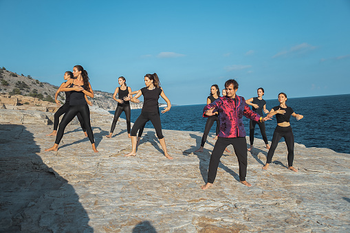 Group of young women doing Tai Chi with instructor on flat stone beach, outdoor exercise on clear summer day