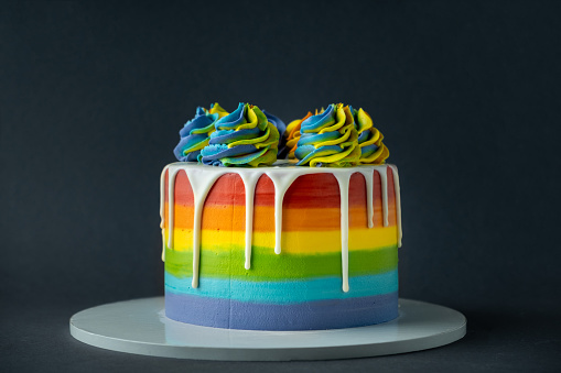 Rainbow cake with whipped cream top on the dark grey background. Birthday cake with multicolored cream cheese frosting and white chocolate drips.