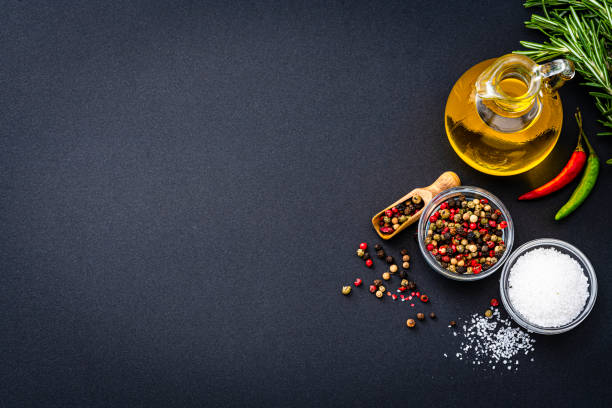 Cooking and seasoning: overhead view of an extra virgin olive oil bottle and two bowls with peppercorns and salt arranged at the right of a black background leaving useful copy space for text and/or logo at the left. High resolution 42Mp studio digital capture taken with Sony A7rII and Sony FE 90mm f2.8 macro G OSS lens