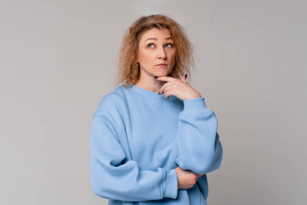 istock Thoughtful middle age woman with curly blonde hair remembering something, standing with doubtful expression in trendy blue sweatshirt over light grey background. Let me think. Doubt concept 1390257423