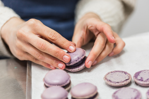 Unrecognizable pastry chef joining lids of purple macaron with filling on table.