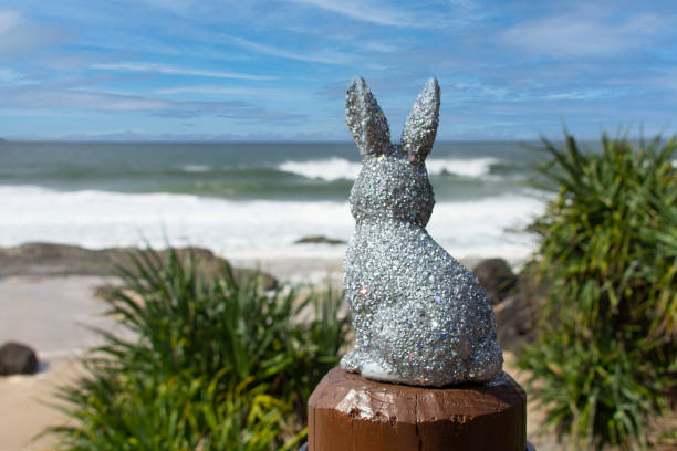 Silver sparkly glitter Easter Bunny at the beach.  Sitting on a brown post looking out to the ocean. Ocean & sky in the background with Pandanus trees down below. Gold Coast Queensland Australia
