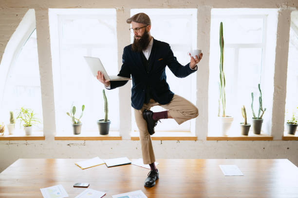 istock Young bearded man, office clerk having fun, doing yoga on wooden table in modern office at work time with gadgets. Concept of business, healthy lifestyle, sport, hobby 1390270295