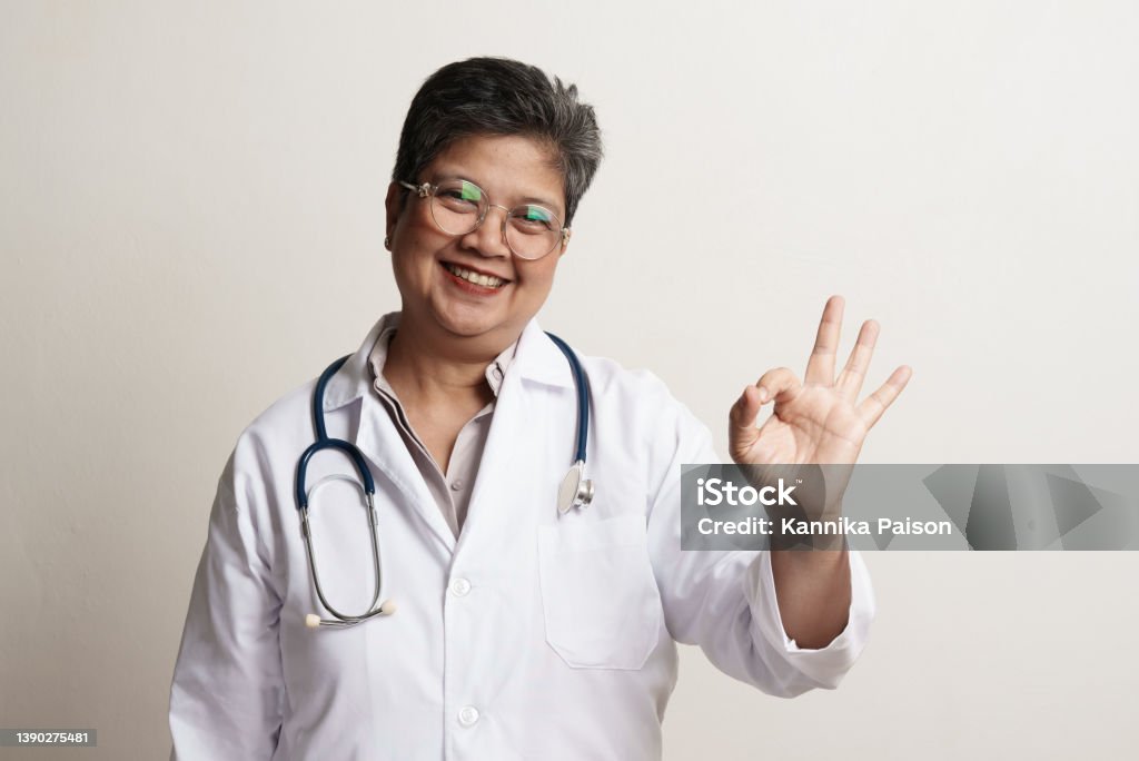 Portrait of confidence senior Asian woman doctor standing against white background Portrait of senior Asian doctor with eyeglasses standing against white background. Female doctor smiling and looking at camera with OK sign. studio portrait advertising banner concept. OK Sign Stock Photo