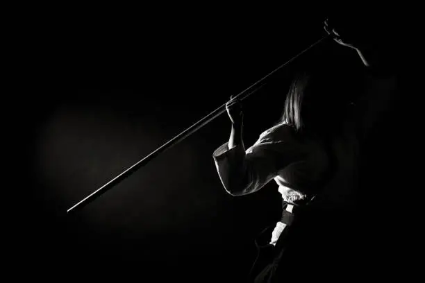 A girl in black hakama standing in fighting pose with wooden sword bokken over dark background. Shallow depth of field. Black and White