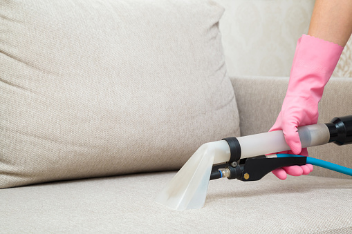 Woman hand in rubber protective glove using spray nozzle of professional vacuum cleaner and washing light beige seat of sofa at home. Extraction method. Commercial cleaning service. Closeup.