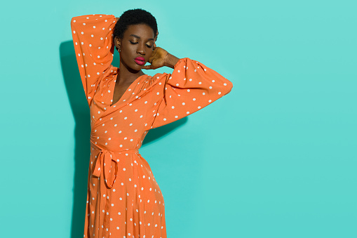 African woman is posing with hands behind head wearing orange long dotted dress. Three quarter length studio shot on turquoise background.