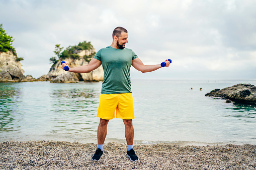 Young cheerful sporty man exercising with dumbbells on the beach