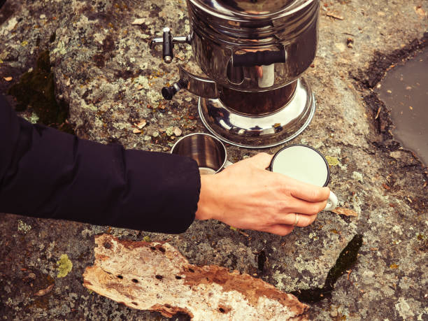 la mano de un hombre coloca una taza de hierro blanco sobre una piedra cerca de un samovar cromado brillante. beber té en la naturaleza en un día de otoño - chromeplated fotografías e imágenes de stock